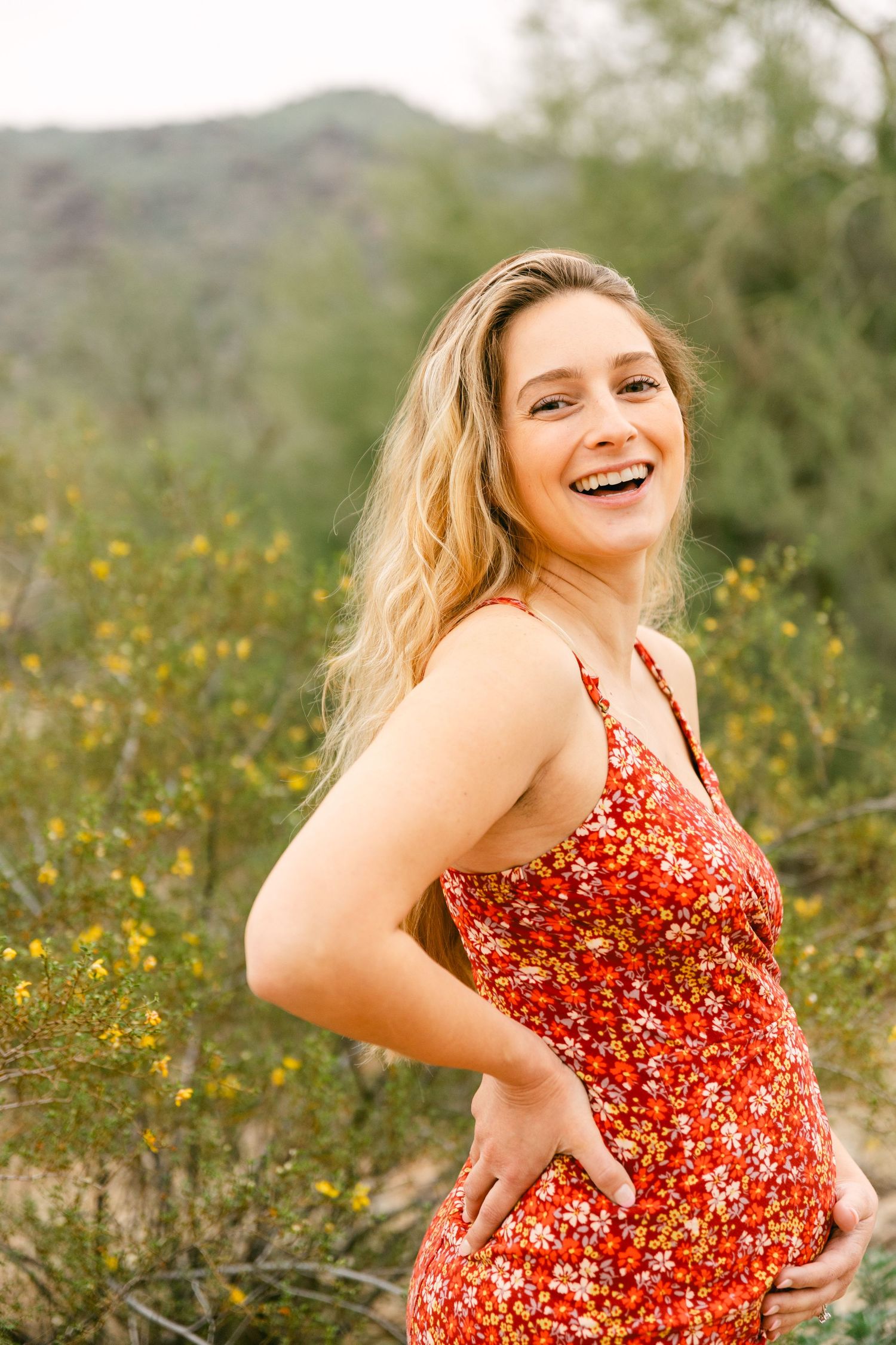 A pregnant woman in a red dress with a hand on her belly and is laughing. There's a green bush with yellow flowers and a mountain behind her