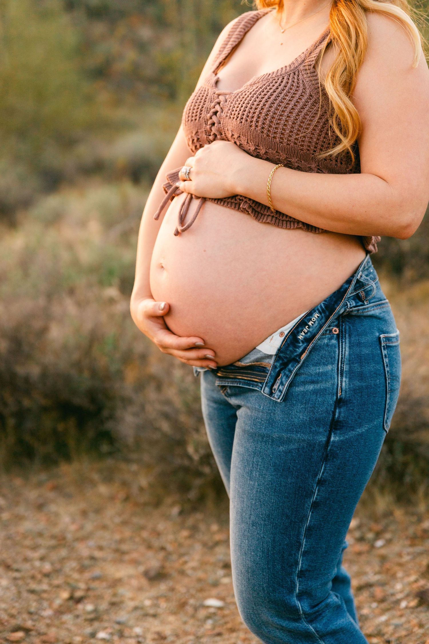 A woman is standing with her hands on her pregnant belly. She's wearing a brown crochet top and a pair of jeans that say "Mom Jeans"