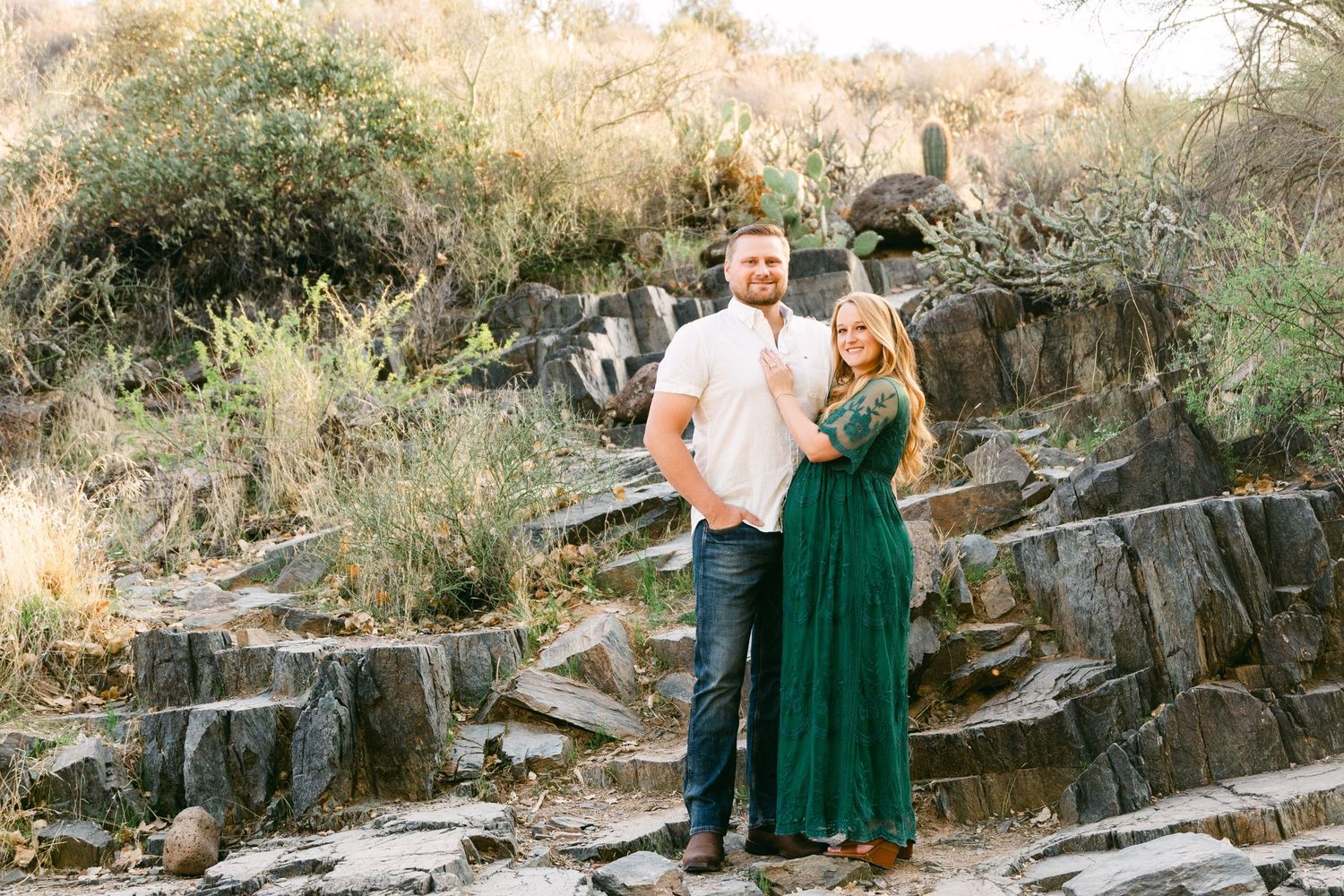 A couple standing together in a rocky area with some greenery. The woman is in a green dress and the man is in a white shirt and jeans.