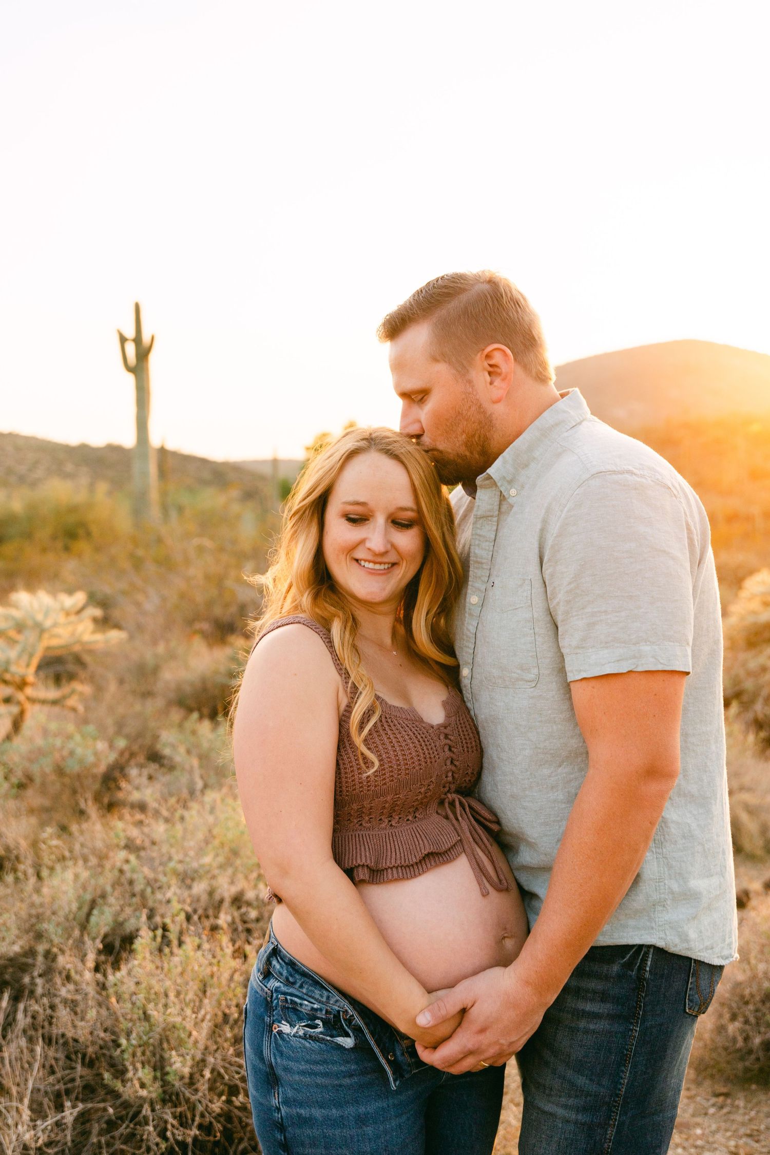 A couple is standing in the desert. He has a hand on her pregnant belly and is kissing her head. She's holding her belly and is looking down