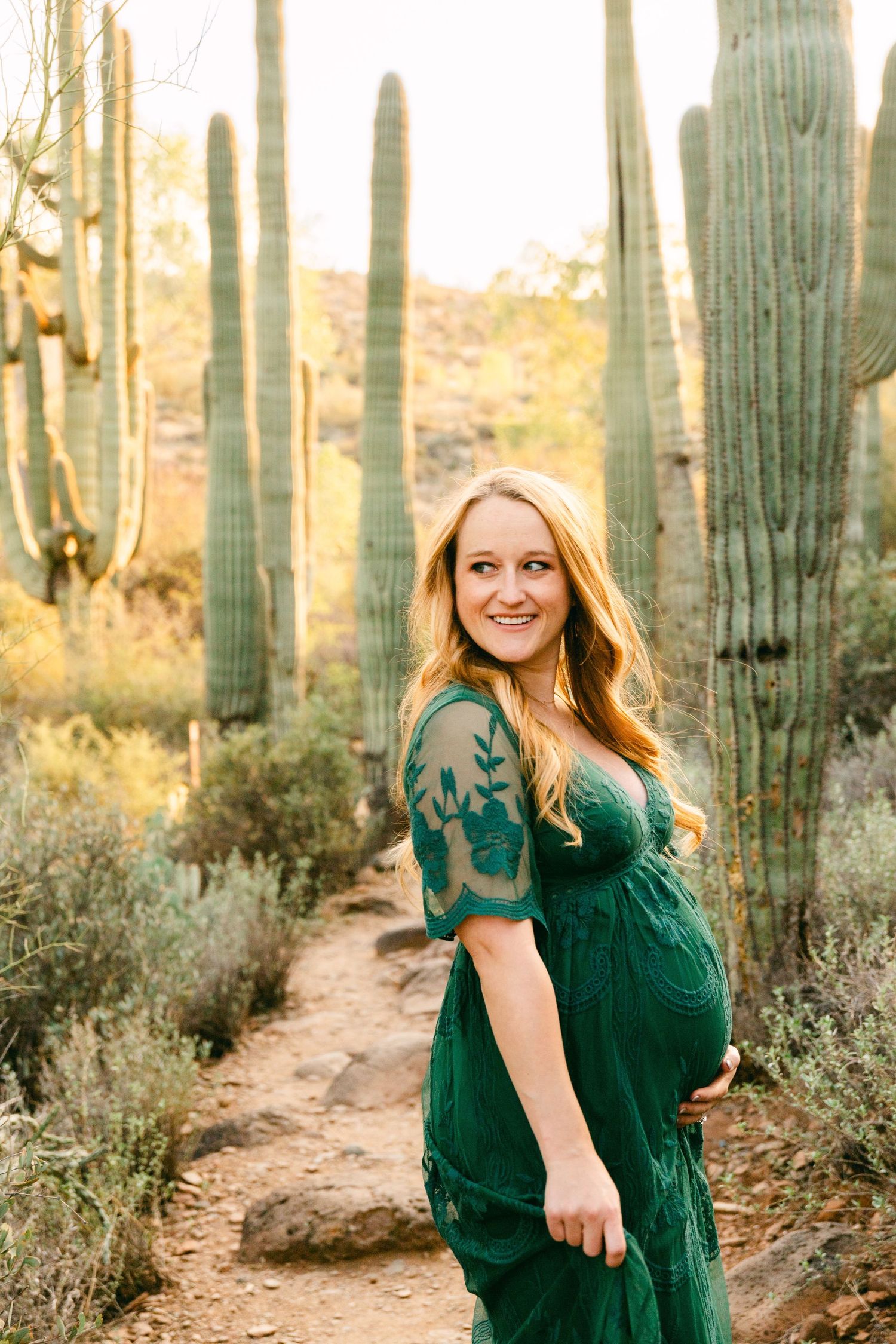 A pregnant woman in a green dress has a hand on her belly and is smiling to the side. She's surrounded by cactus and a rock path.