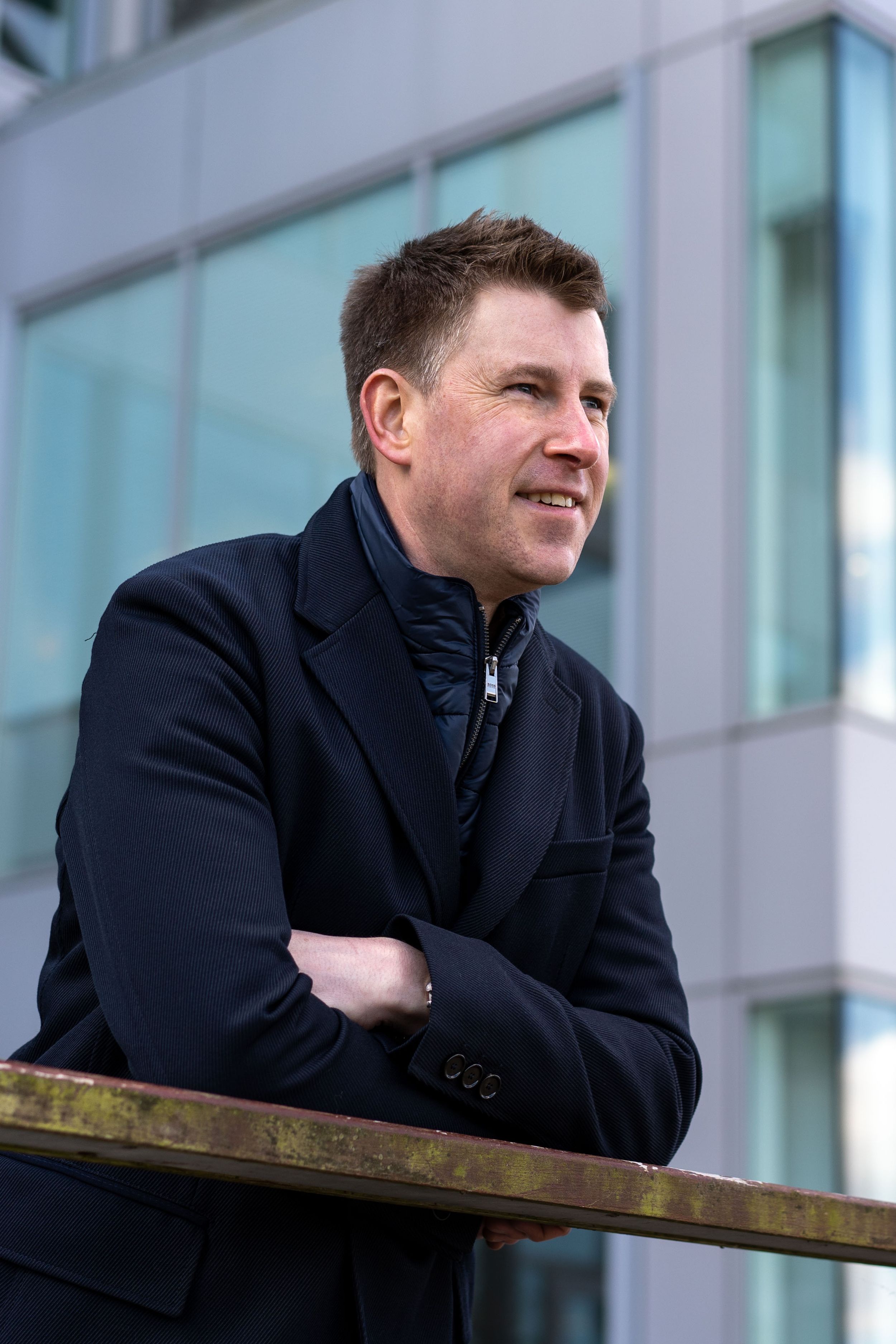Founder in navy coat leans on wooden railing while smiling on personal branding photoshoot in a Whiteley Business Park, Fareham, Hampshire