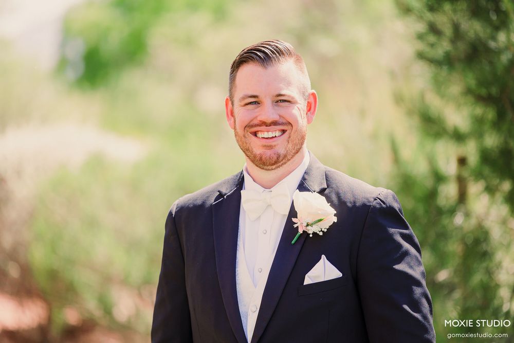 Row of groomsmen in dark suits posing outdoors in a natural garden setting.