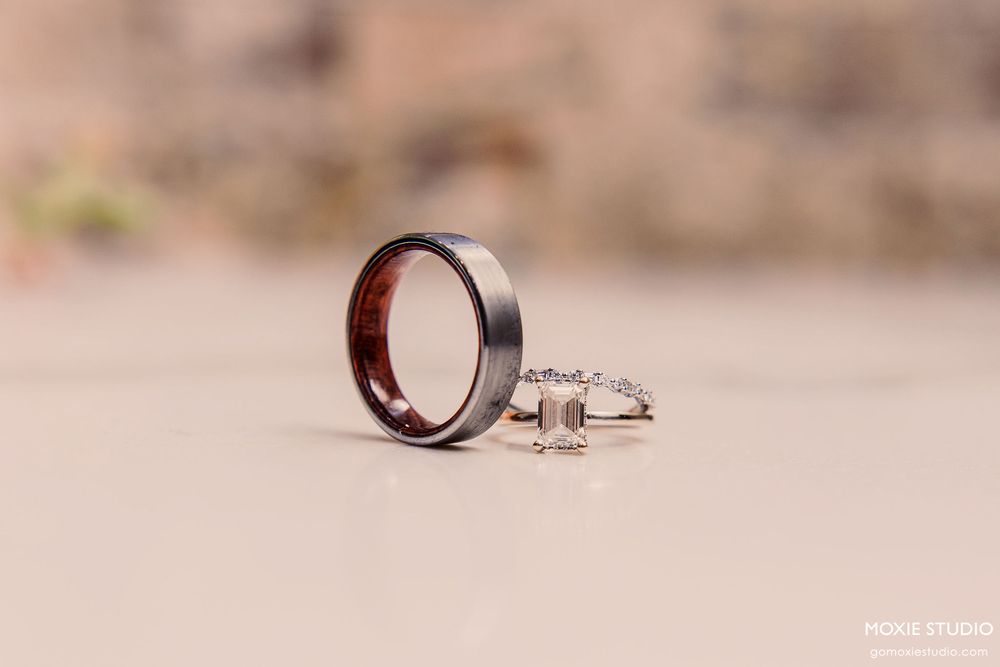 Two wedding rings, one tungsten band and one diamond engagement ring, photographed on a blurred beige background.
