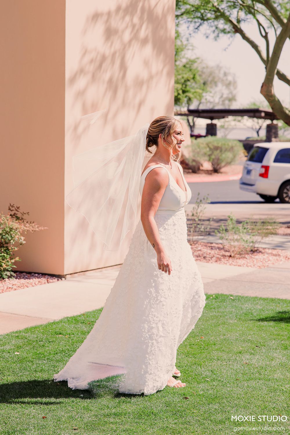 Bride in white lace dress walking gracefully on green lawn near stucco building.