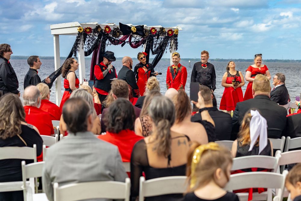 Beachfront wedding ceremony with black draped arch, guests in red and black attire by the water.