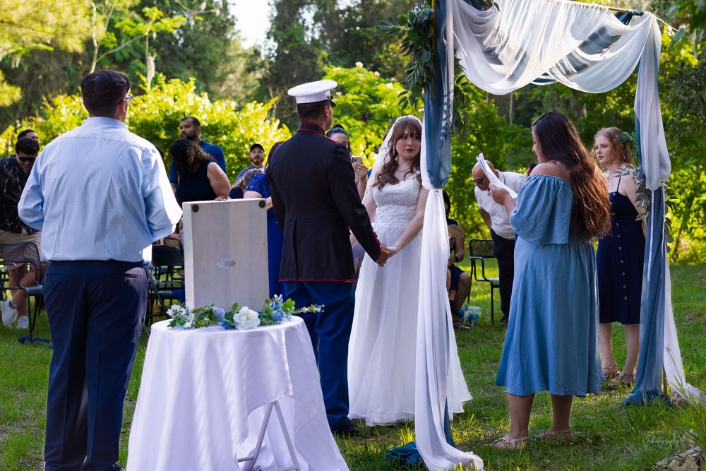 Outdoor military wedding ceremony with bride in white cape gown and groom in dress uniform under draped arch.