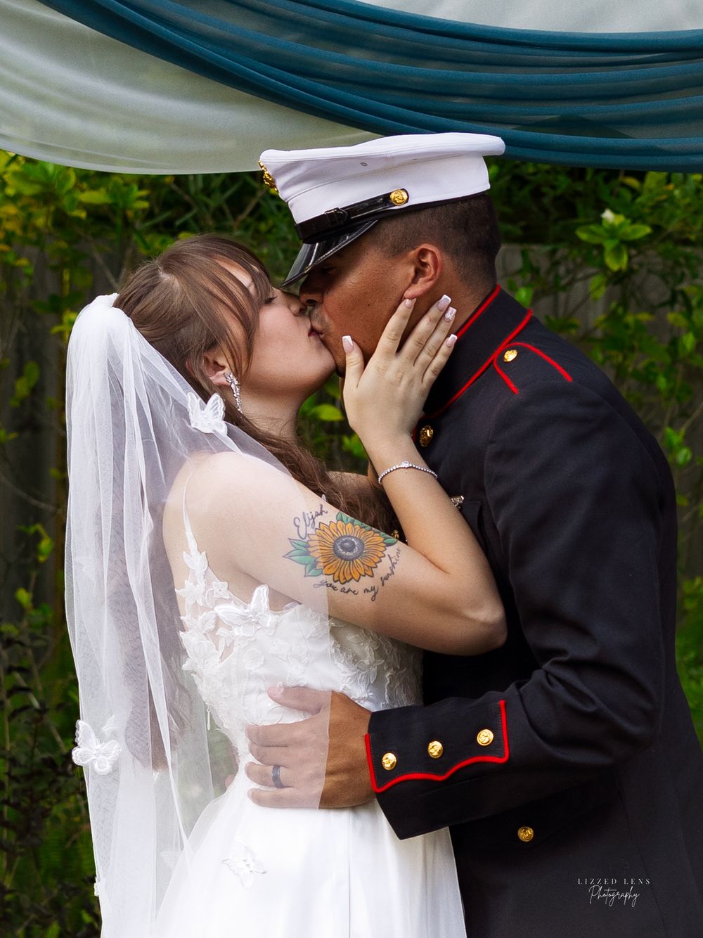 Close-up of tattooed bride and Marine groom sharing passionate first kiss under teal wedding arch.