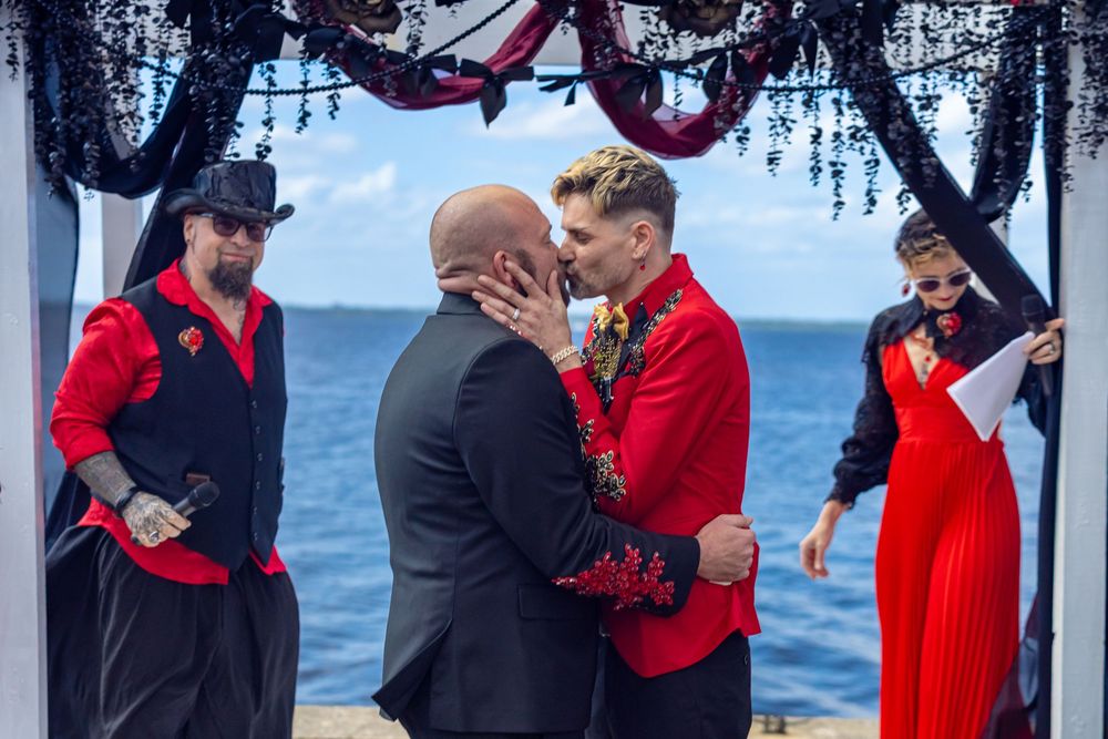 Two grooms share first kiss at outdoor beachfront wedding ceremony with wedding party.