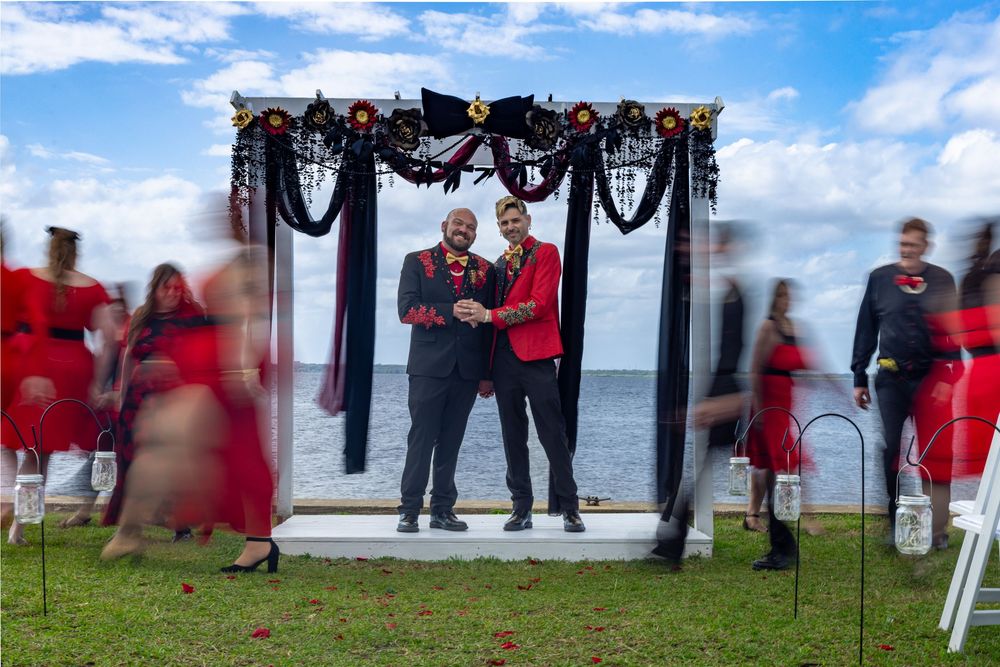 Two grooms stand at a decorated outdoor altar by the water surrounded by guests in red.