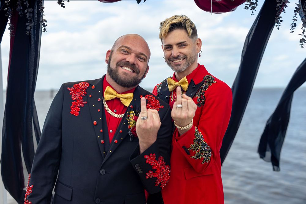 Two grooms in matching red and black floral suits show wedding rings while smiling by the waterfront.