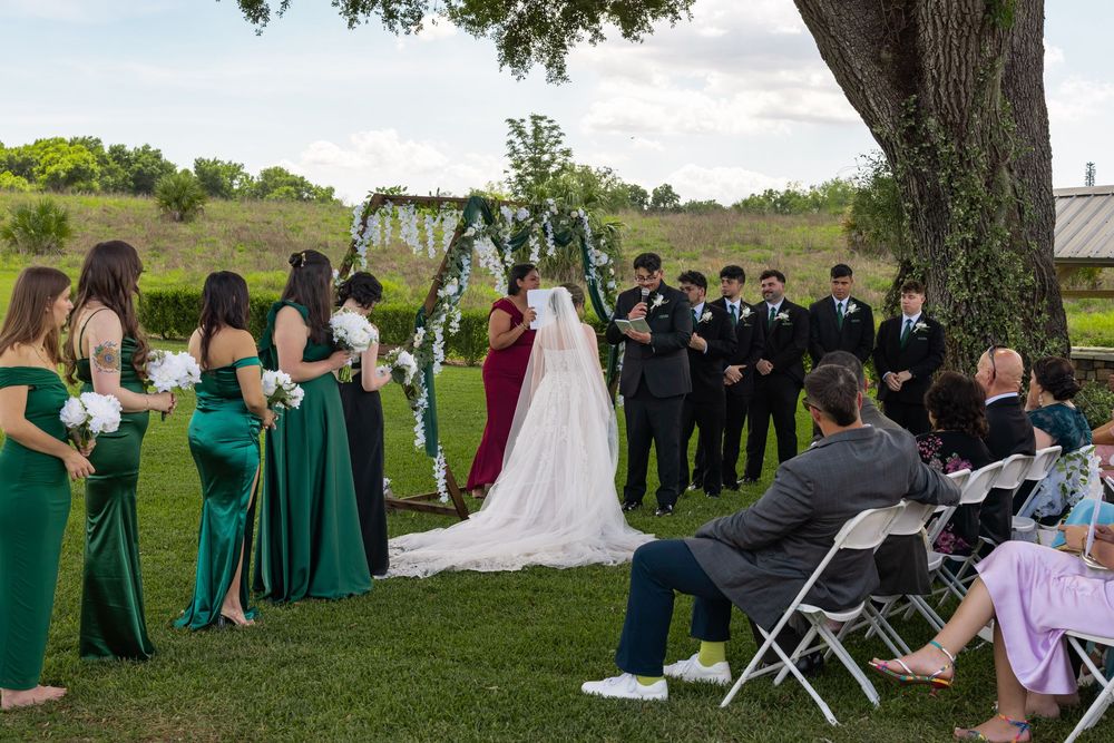 Wedding ceremony outdoors with green bridesmaid dresses and groomsmen in black suits under a floral arch.