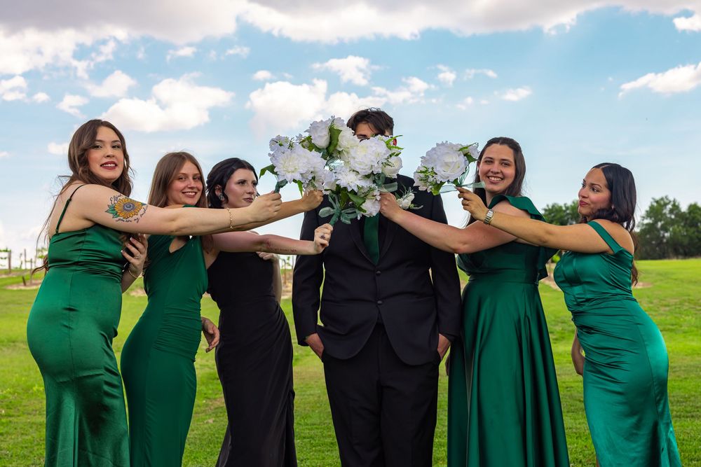 Groom surrounded by five bridesmaids in emerald green dresses holding white bouquet outdoors.