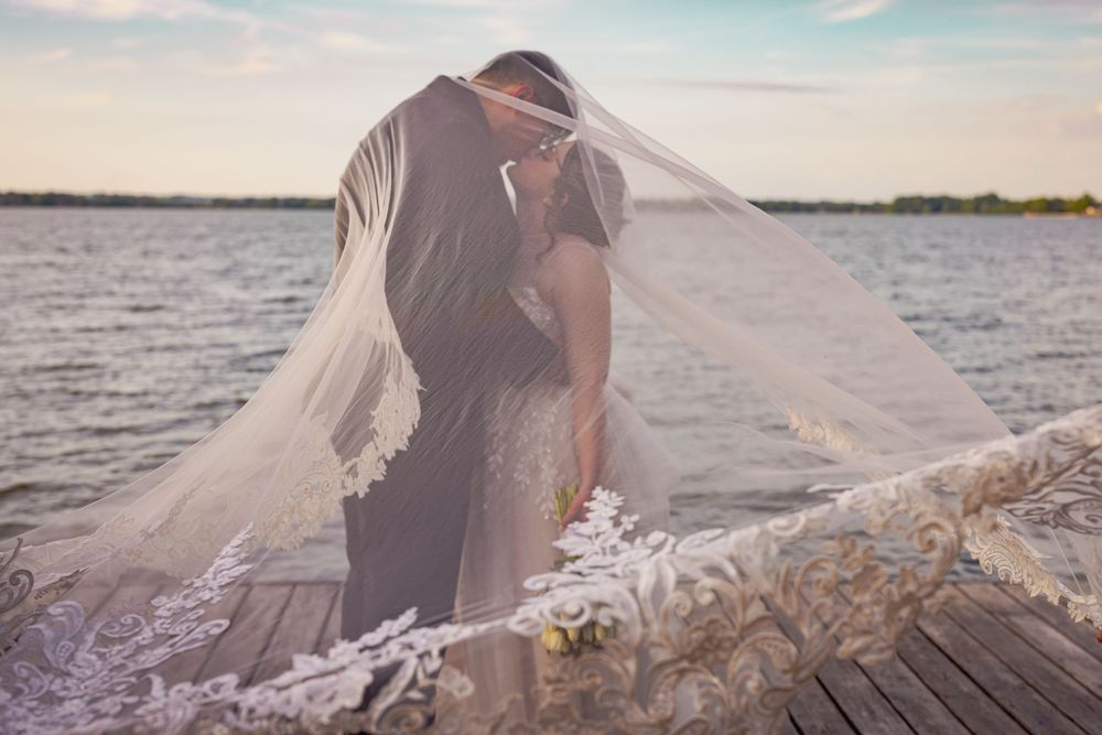 Bride and groom share a kiss under flowing wedding veil on a lakeside dock at golden hour.