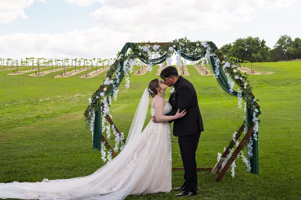 Bride and groom share tender kiss under floral wedding arch on scenic green vineyard lawn.