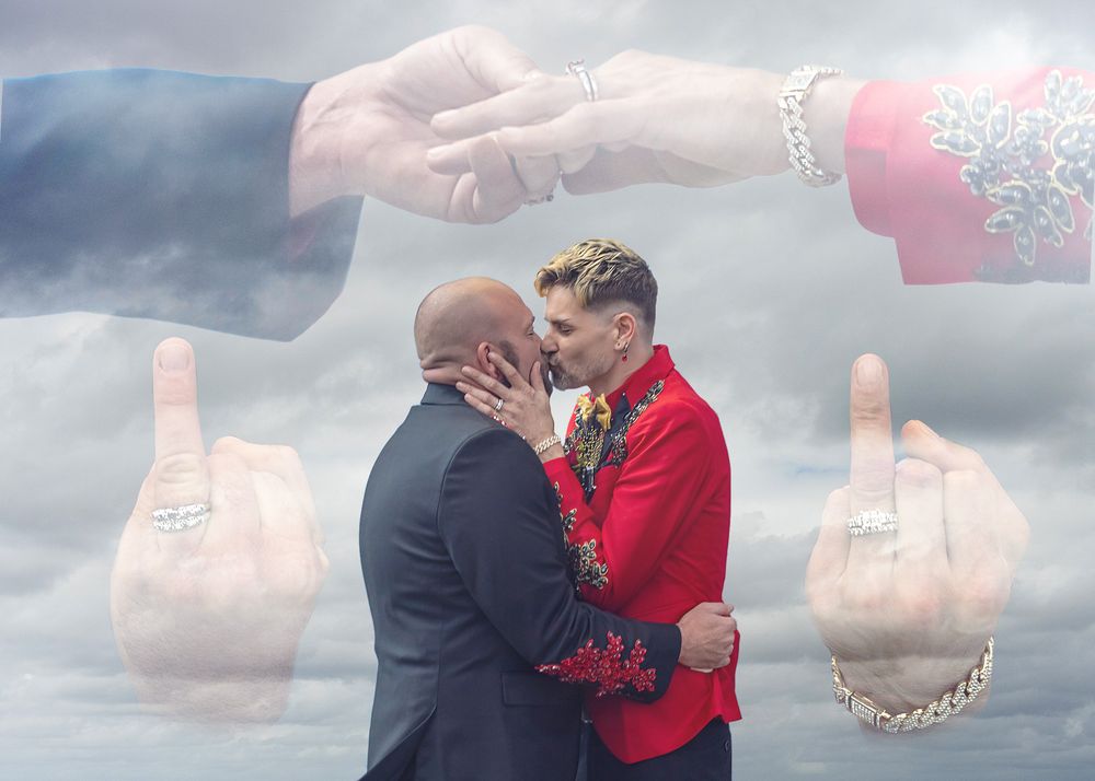 Two grooms kiss at their wedding with artistic hand overlays in the cloudy sky.