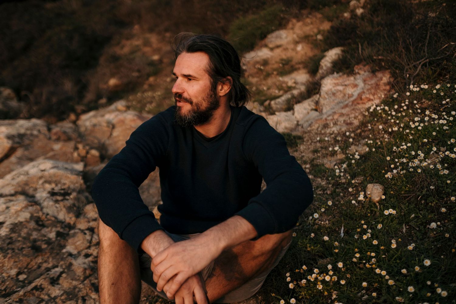 Person in black sweater sits thoughtfully among rocks and wildflowers during golden hour.