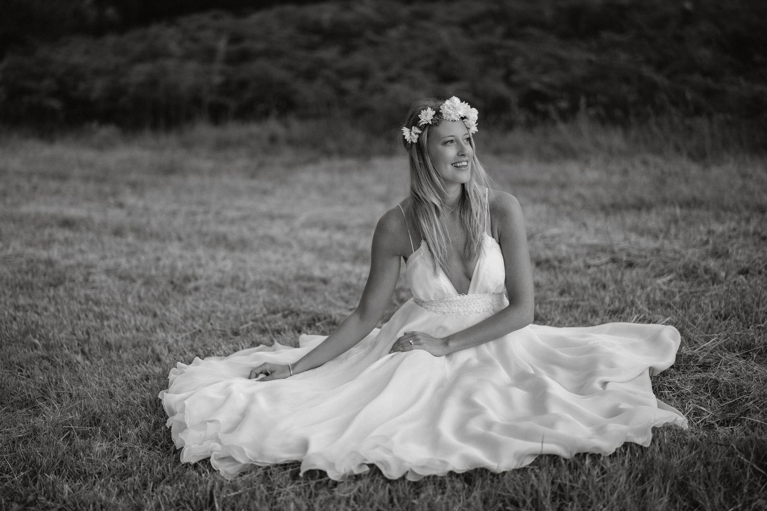 Black and white portrait of someone in a flowing white dress and floral crown sitting gracefully in a meadow.