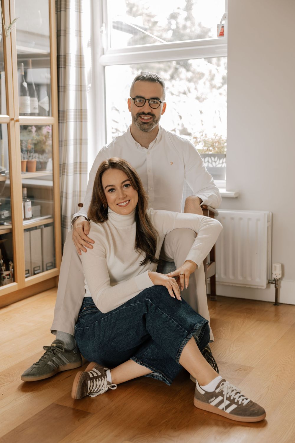 A couple in matching white sweaters sits casually by a bright window on hardwood floors in a cozy home setting.