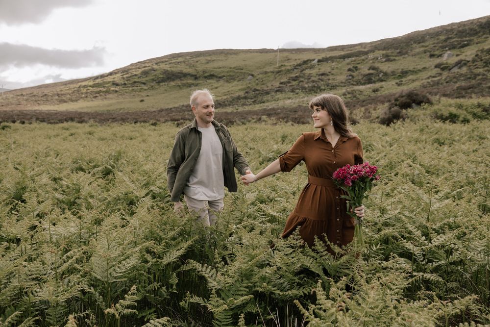 Young couple runs joyfully through tall ferns in scenic countryside under moody sky in Ireland.