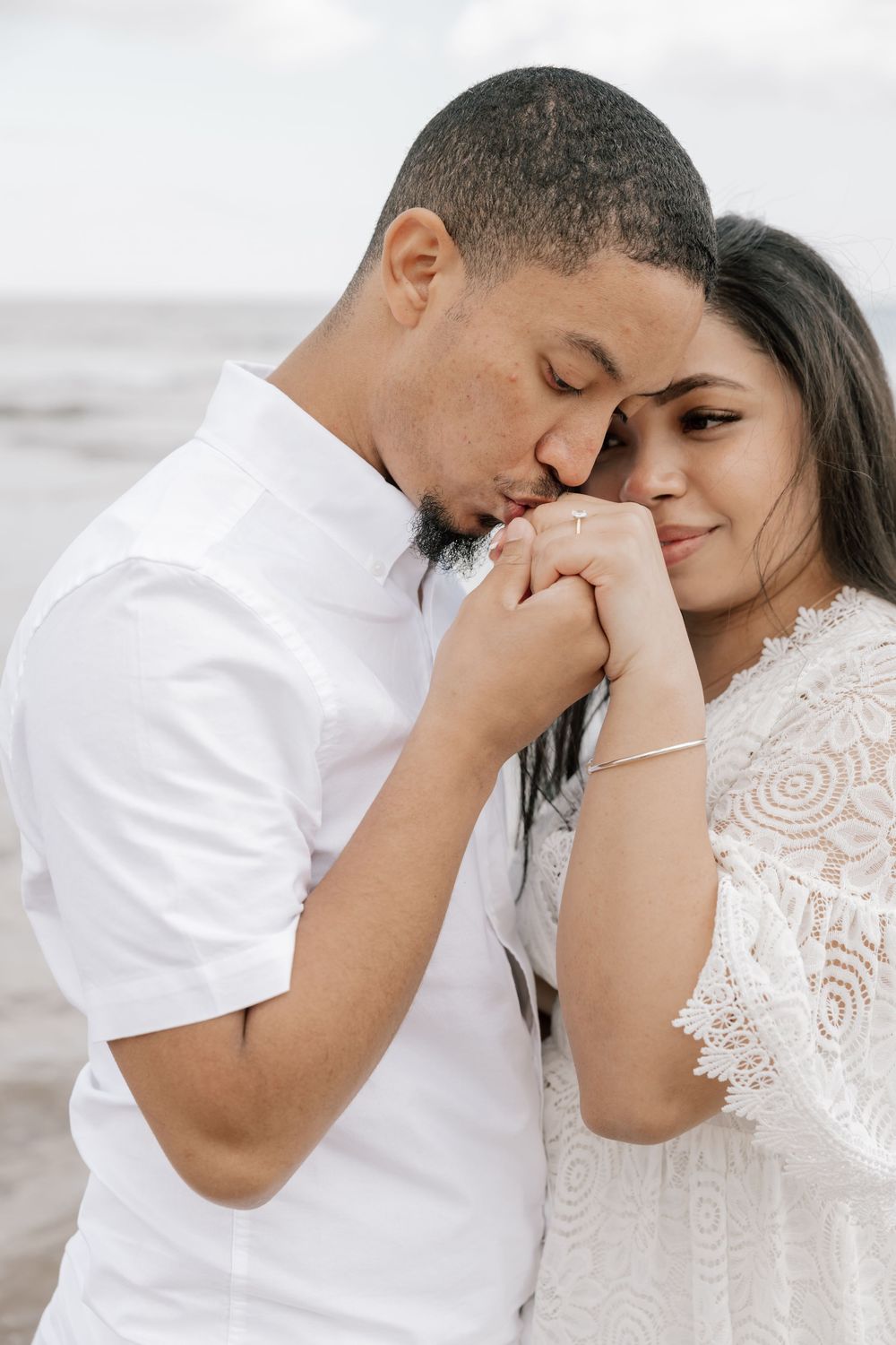 Romantic couple embraces on beach, with groom kissing bride's hand as she wears delicate lace dress.