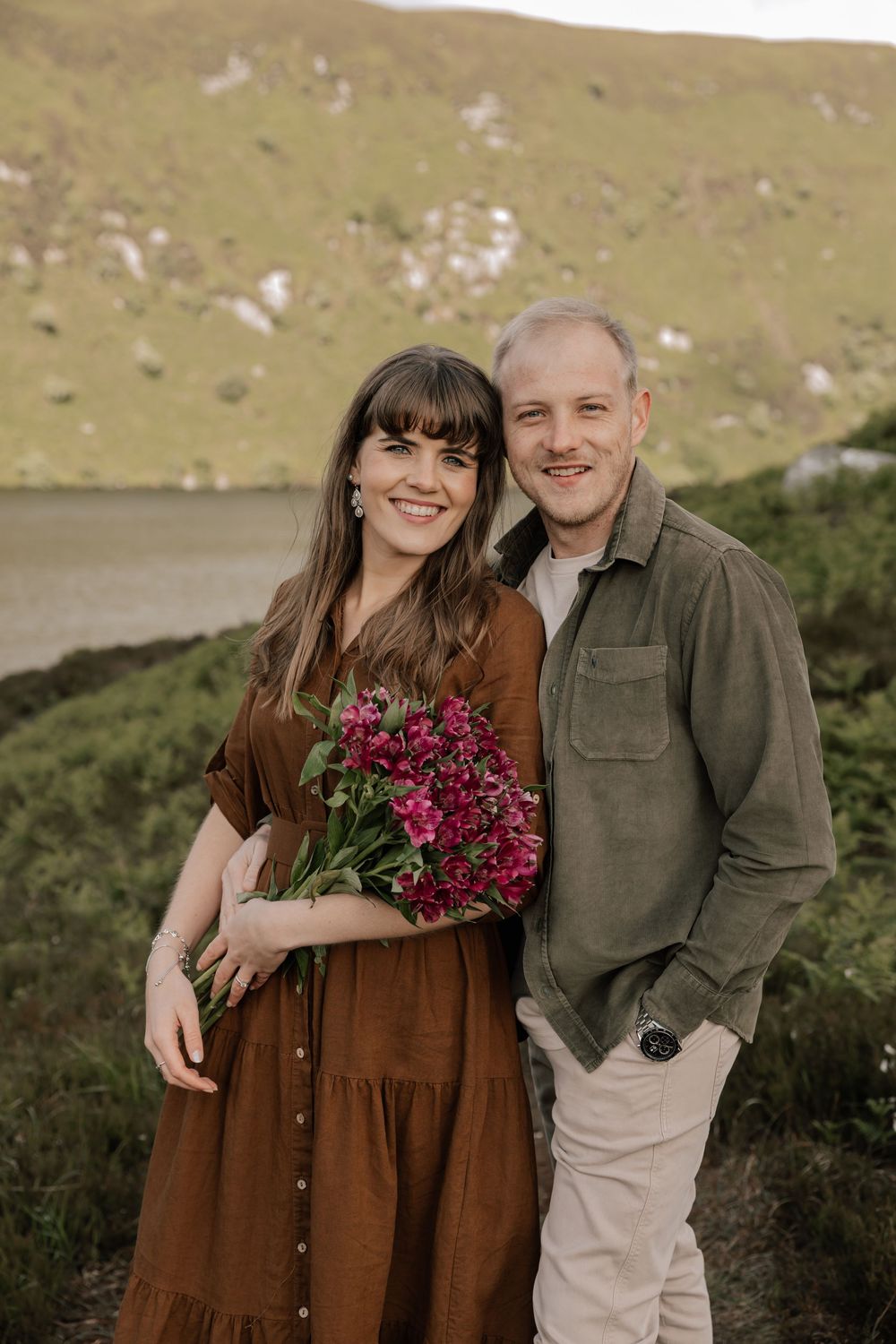 A couple stands together on a hillside by a lake, with the person in brown dress holding pink flowers against scenic landscape.