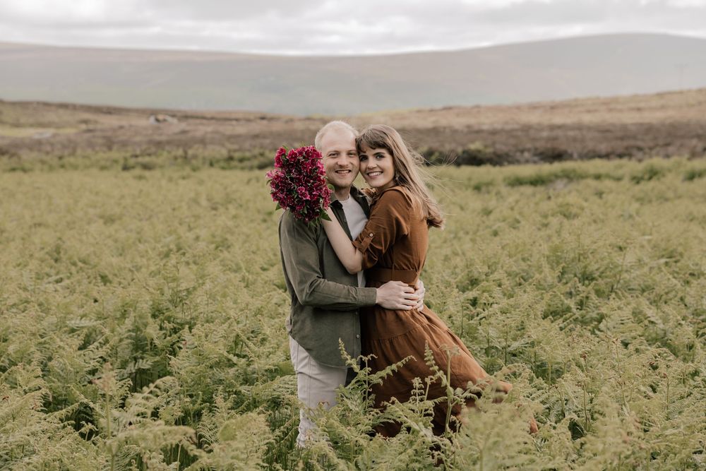 A couple embraces while standing in a vast green field under an overcast sky during autumn.