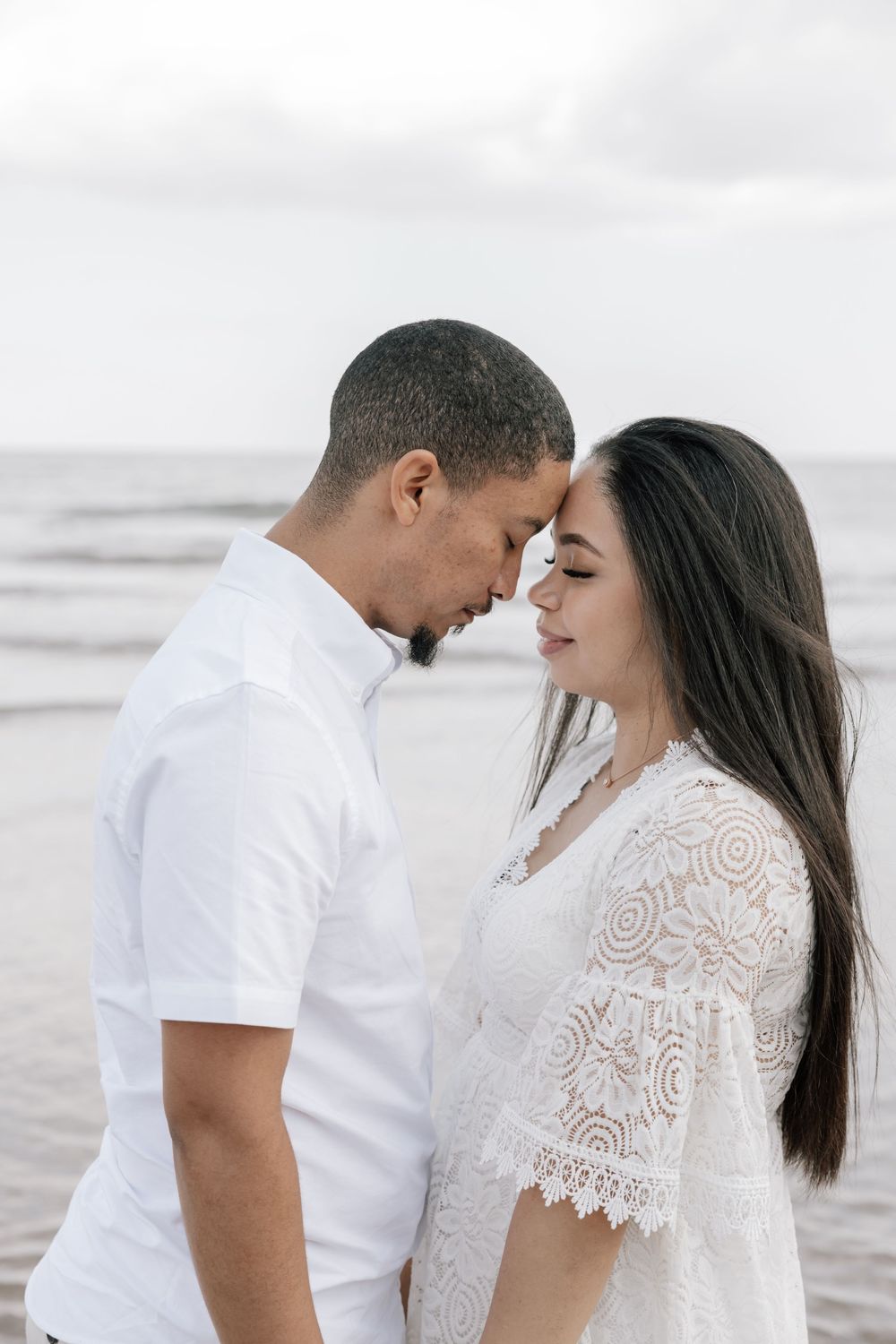 Romantic couple in white outfits sharing an intimate moment on a cloudy beach.