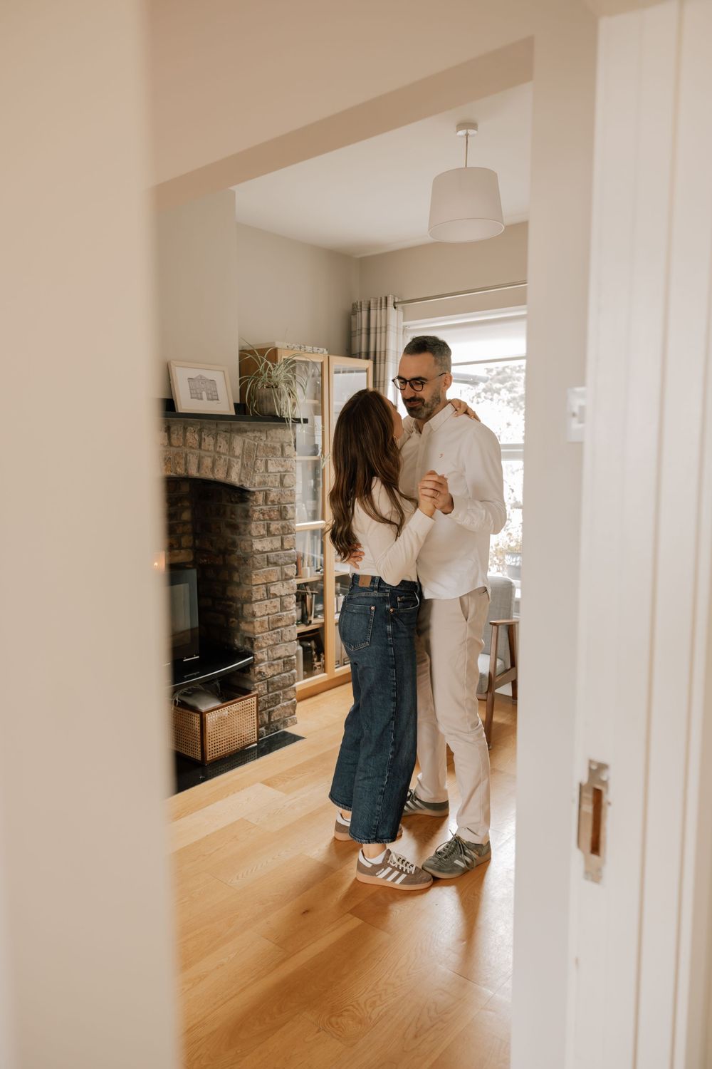 A couple embraces in a cozy living room with hardwood floors and a stone fireplace.