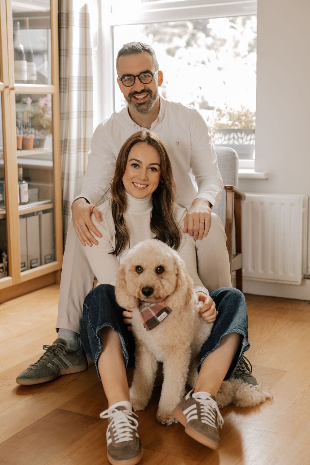 A couple sits at home with their fluffy cream-colored Goldendoodle dog on a hardwood floor by a window.