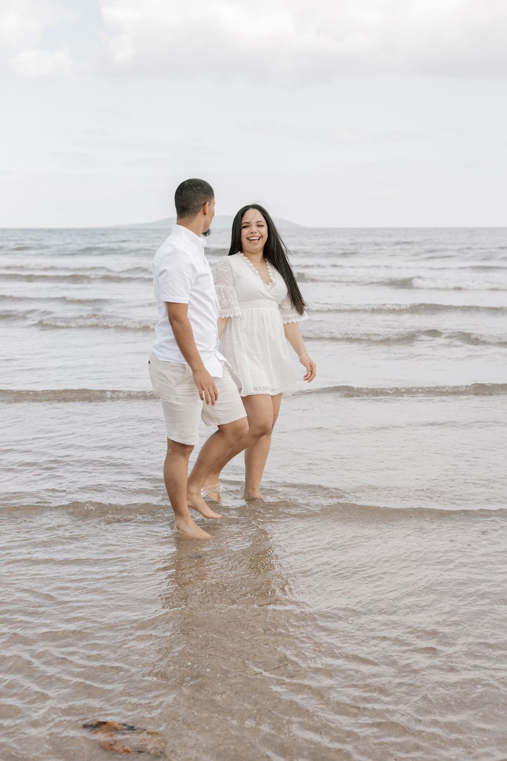A couple in white outfits stands together in shallow ocean waves on a cloudy beach day.