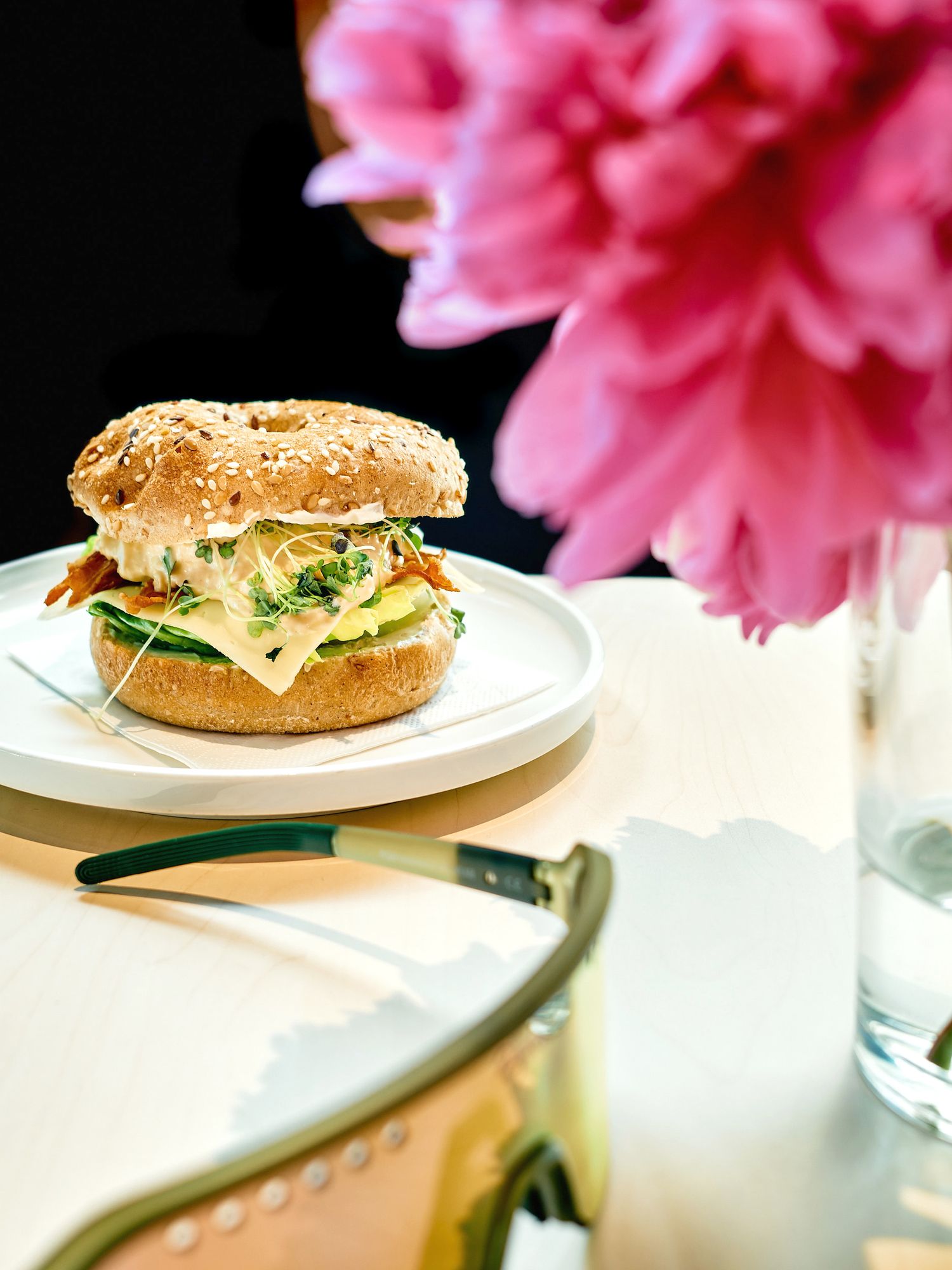 A gourmet burger with cheese and lettuce on a sesame seed bun sits on a white plate beside pink flowers and glassware.