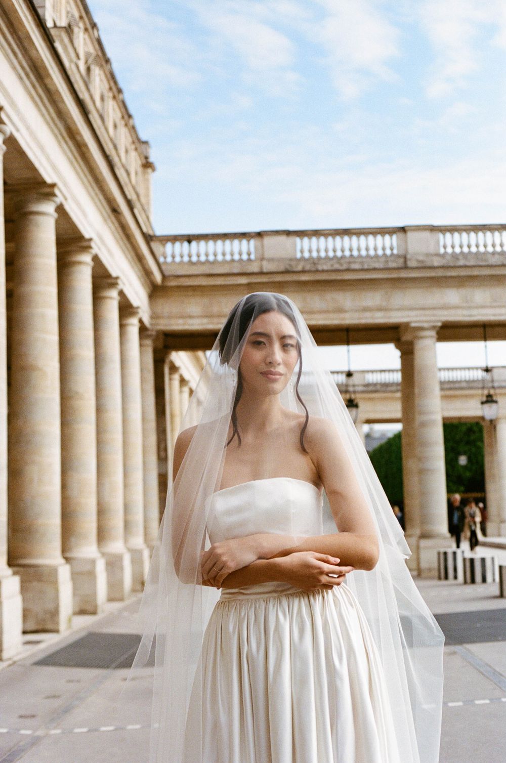 Elegant bride in white strapless wedding gown and veil poses at classical colonnade with stone pillars and balustrade.
