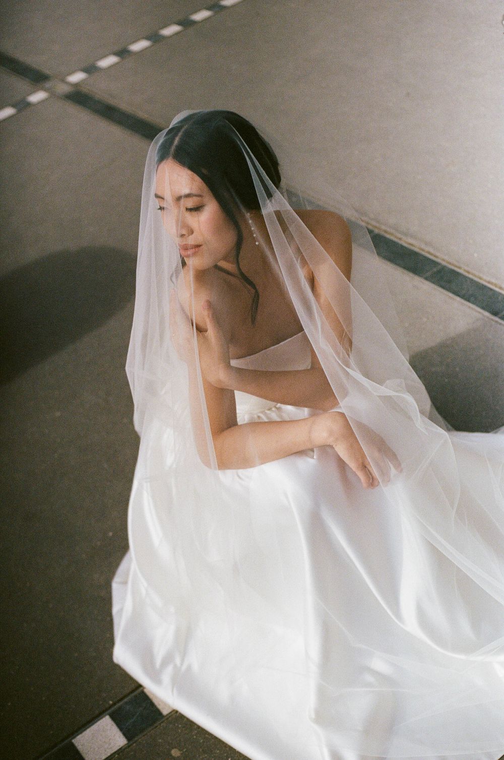 Bride wearing elegant white dress and flowing veil sits thoughtfully in minimalist indoor setting with dramatic lighting.