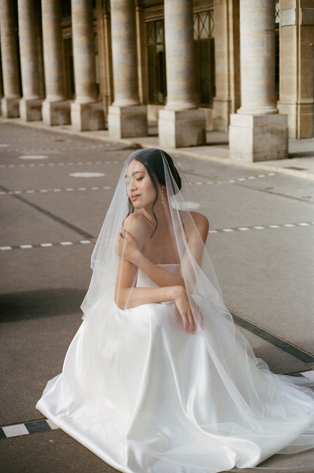 A bride in a flowing white wedding gown and veil poses elegantly among classic stone columns.