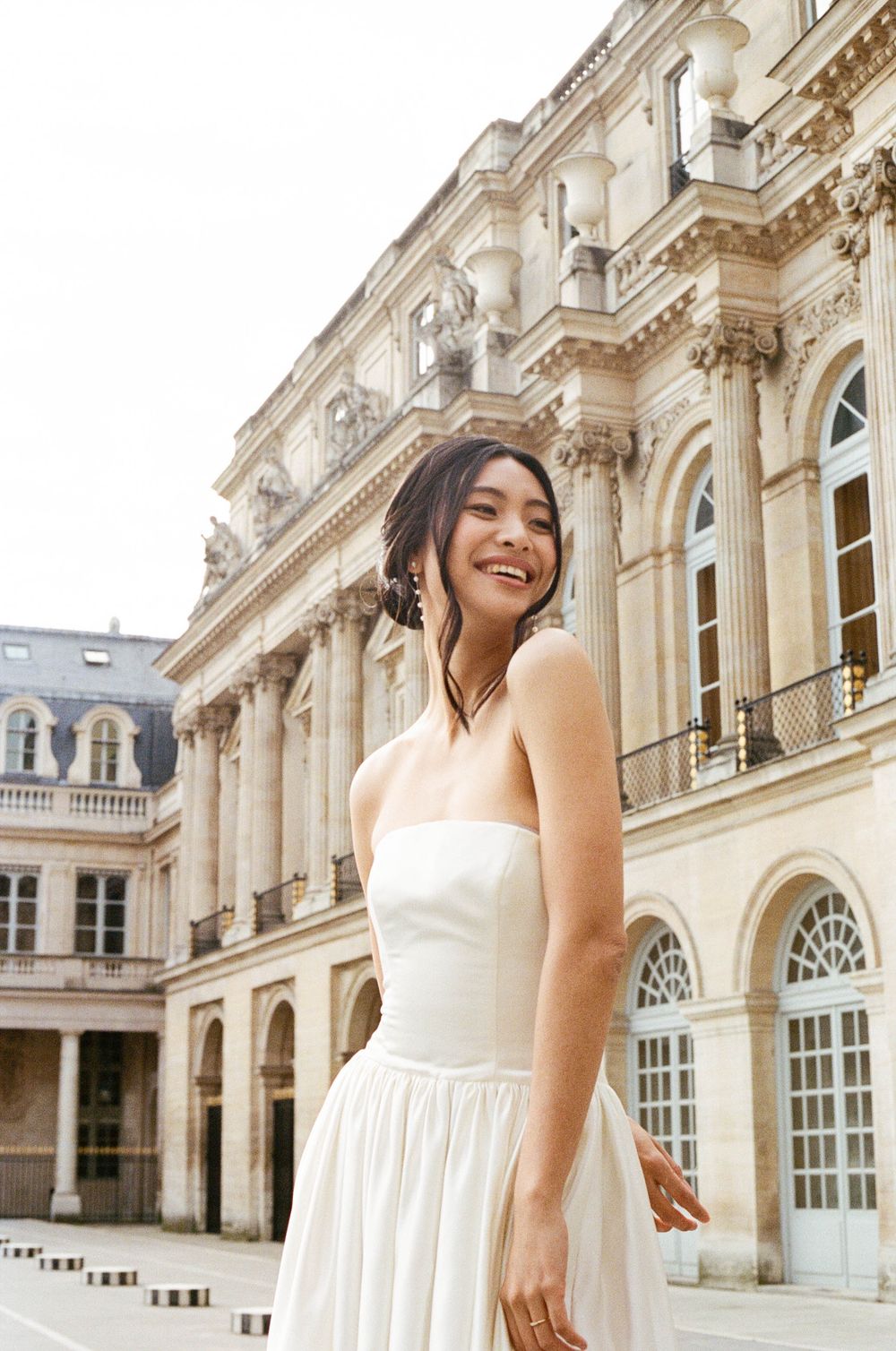 A bride in a strapless white wedding gown standing at an elegant French palace with ornate architecture.