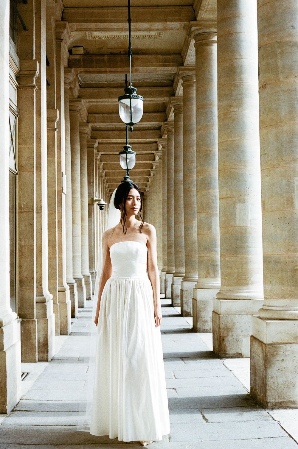 A white wedding dress stands out against classical stone columns and hanging lanterns in a grand colonnade.