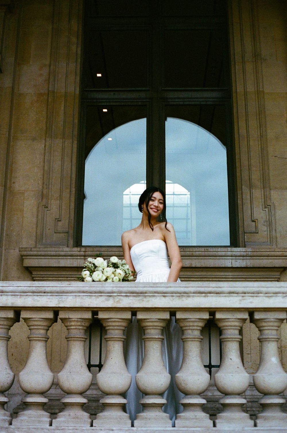 Bride in a white strapless gown stands at ornate stone balcony with white roses.
