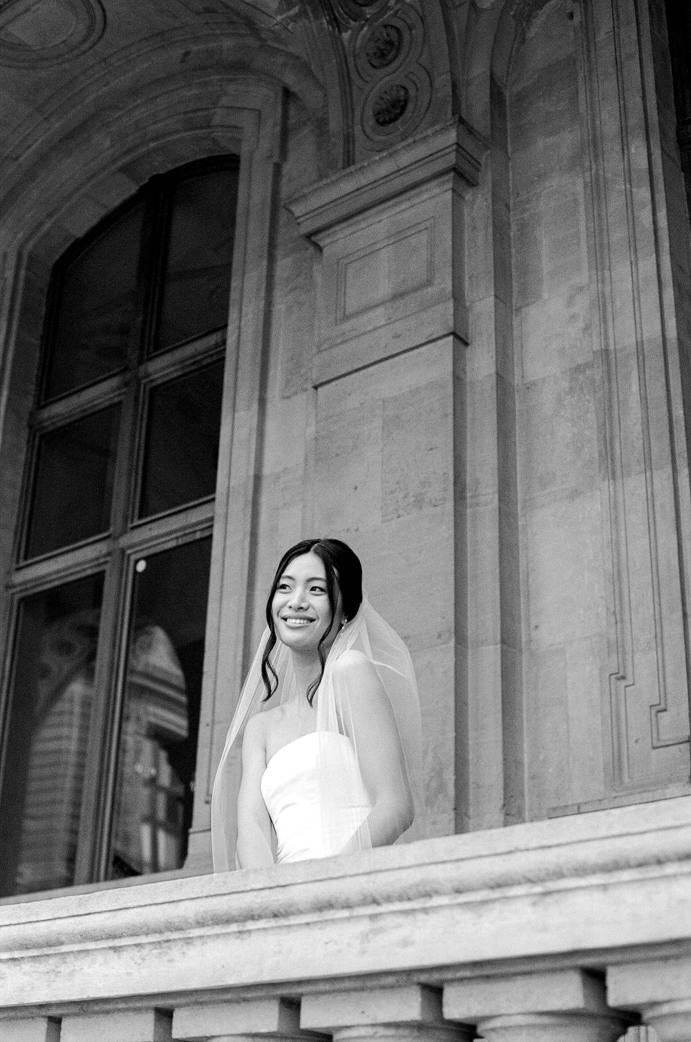 A black and white portrait of a bride in a wedding dress and veil standing by an ornate architectural window.