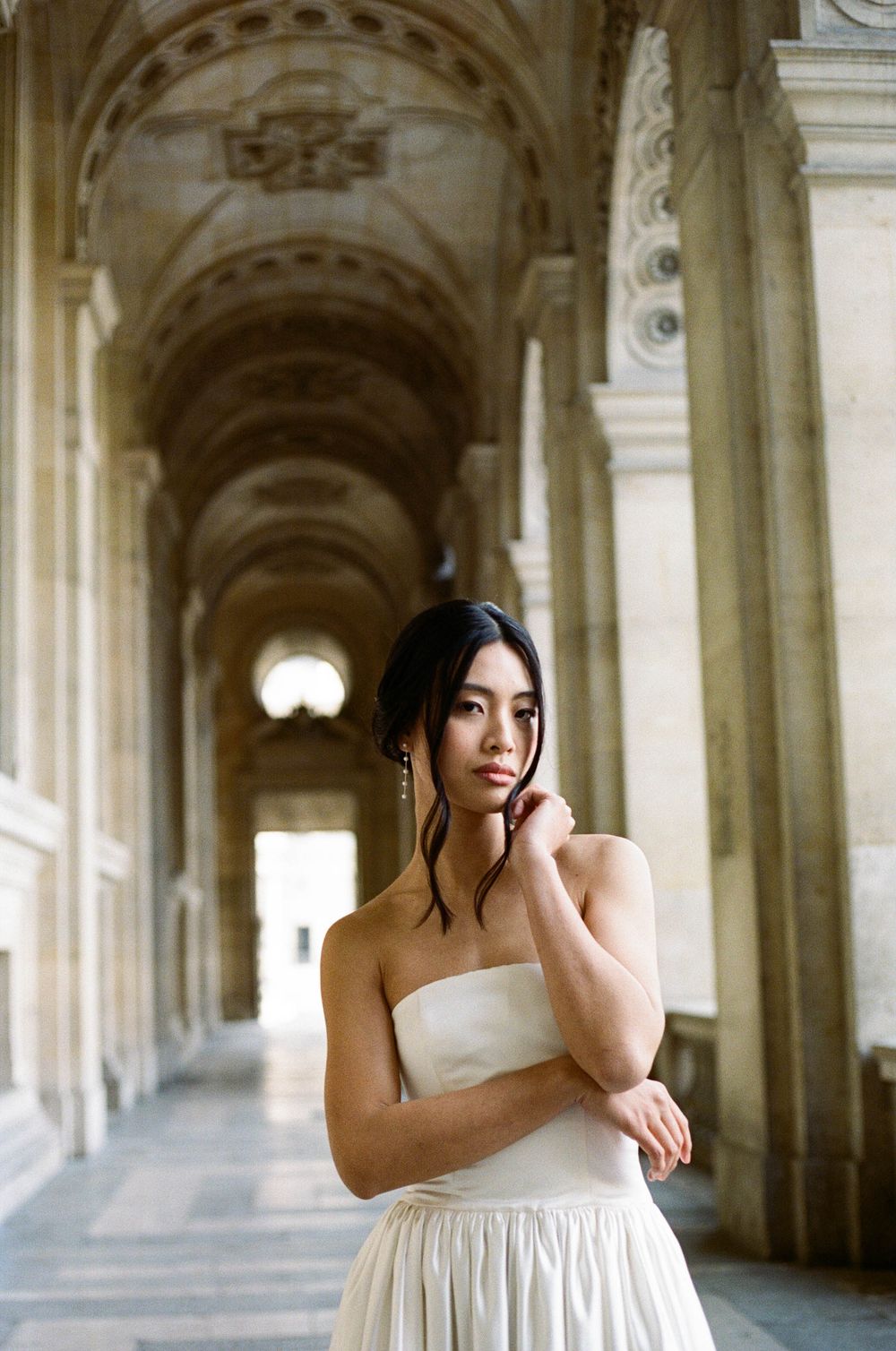 A figure in an elegant white gown poses thoughtfully in an ornate columned hallway with vaulted ceilings.