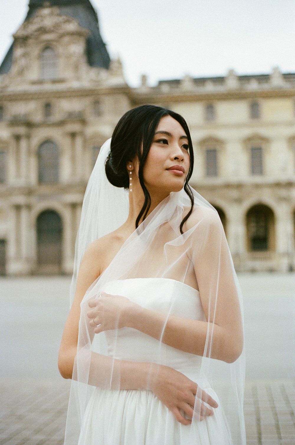 Elegant bride in white strapless gown and sheer veil poses outside historic European building with classical architecture.