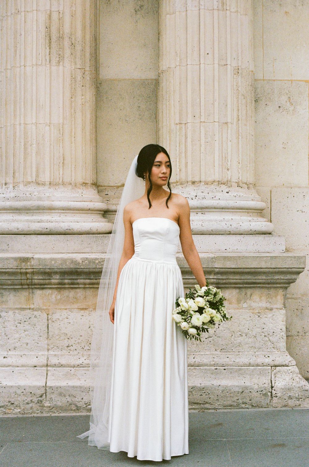 A bride in a classic white strapless wedding gown holds a white rose bouquet against classic stone columns.
