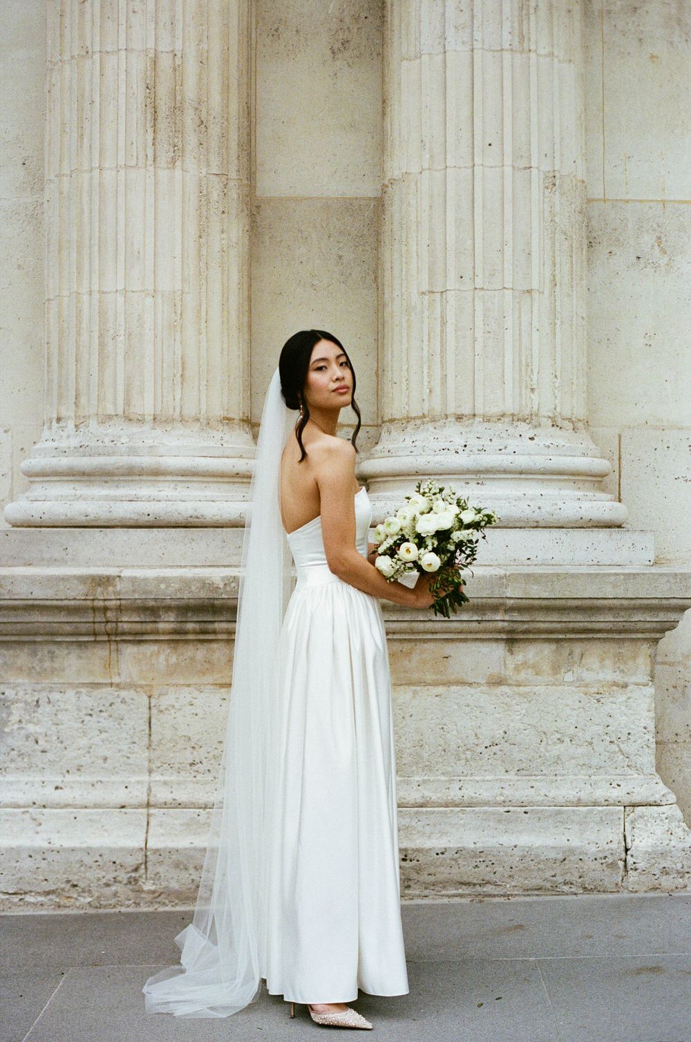A bride in a flowing white dress holds a bouquet of white flowers while standing against classical stone columns.