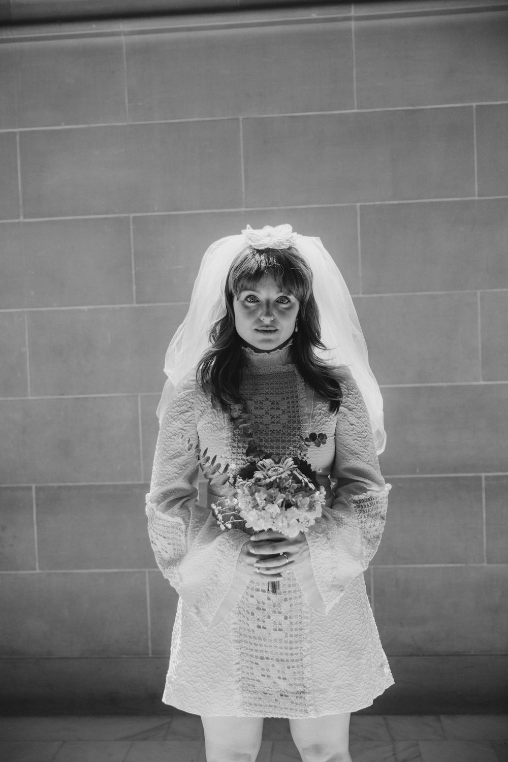 A bride in a short white dress holds a bouquet against a stone wall.