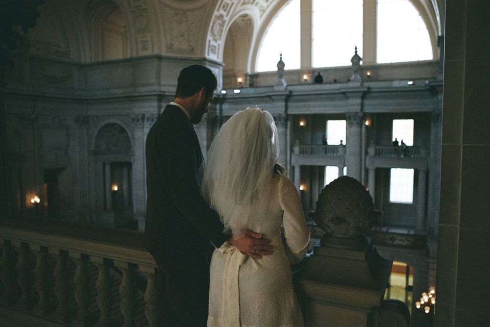 A couple stands at an elegant balcony overlooking an ornate interior.