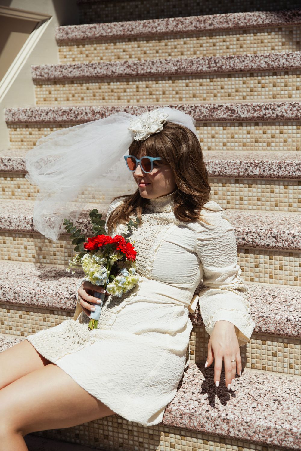 Bride in short white dress and sunglasses sitting on mosaic steps with red bouquet.