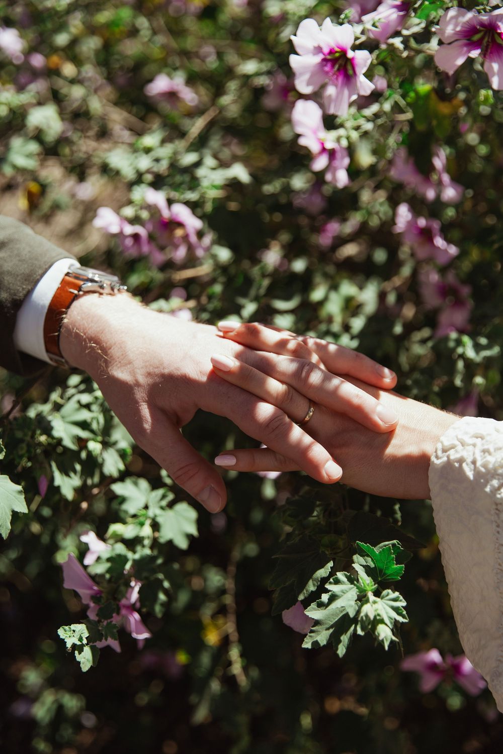 Close-up of hands touching with wedding rings against purple flowers in garden.