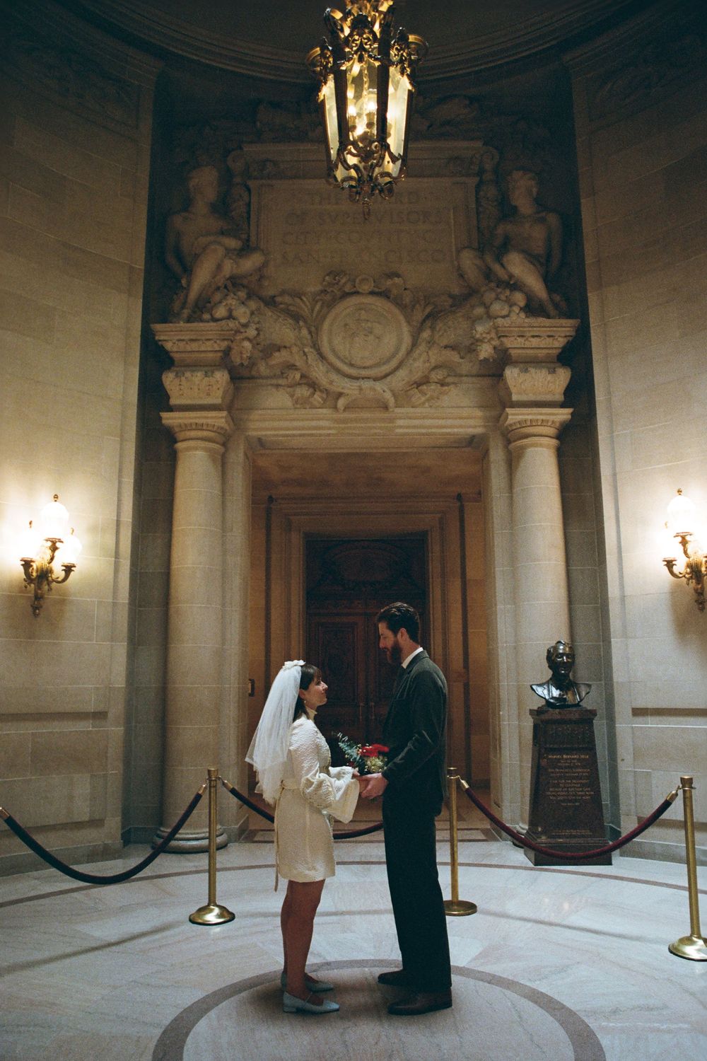 A couple stands in a grand marble rotunda with classical columns and ornate architectural details.