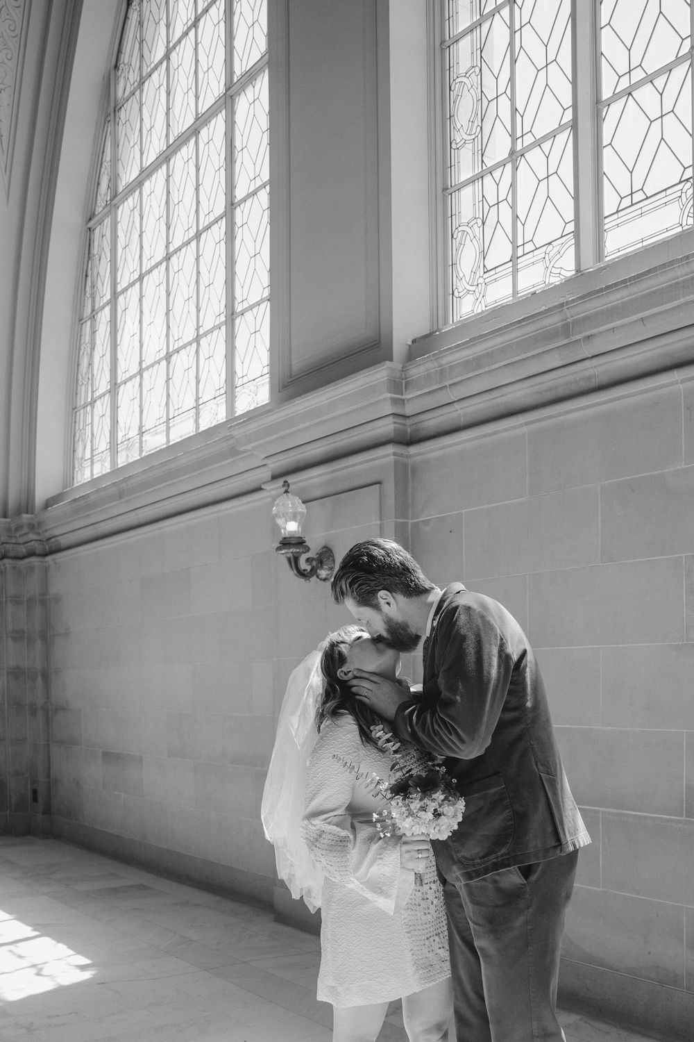 A tender moment between a couple in formal attire near tall windows.