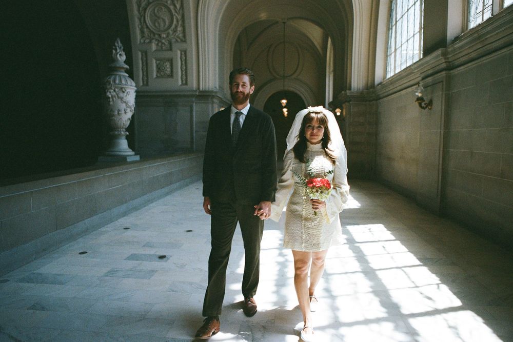 A couple walks through a grand hallway with sunlight streaming through windows.